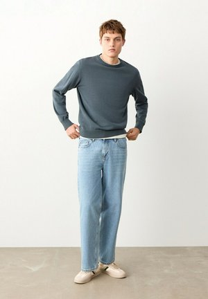 Mladi moški, oblečen v temnosivo jopico, svetlo modre kavbojke in bele superge, stoji v notranjosti ob beli steni z rokami blizu pasu.