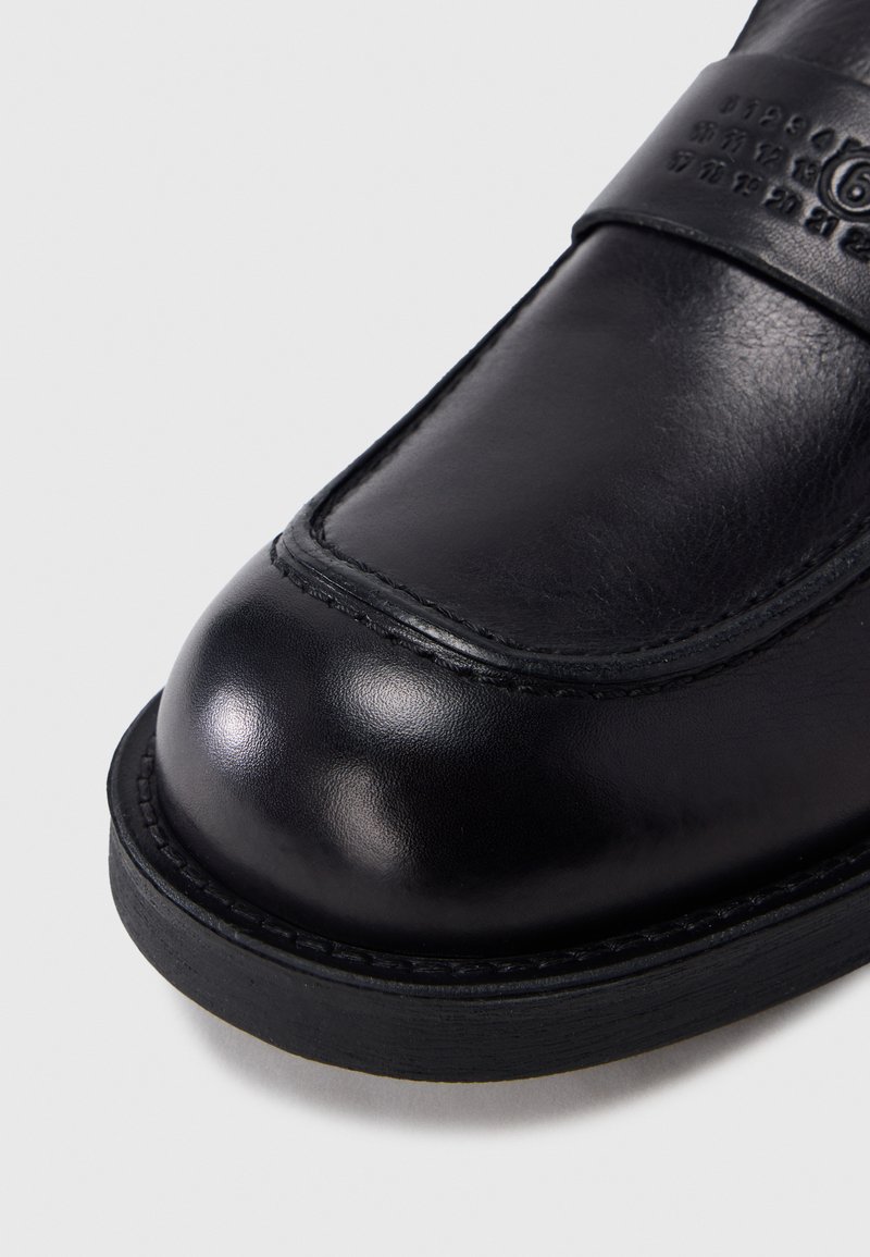 Sort lædersko med rundet, glat afslutning og præget tal på oversiden nær snuden, hvilende på flad sort sål.