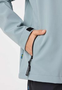 Hand in light blue jacket pocket with zipper pull labeled "SWEDEMOUNT" and Swedish flag tag on jacket side seam.