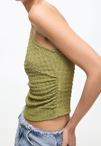 Haut sans manches vert olive avec un design texturé et froncé. Coupe ajustée avec des détails froncés sur le côté. Associé à un short en denim effiloché.