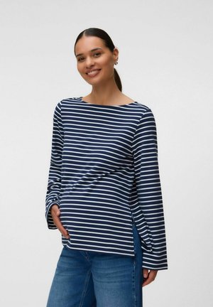 Femme enceinte souriante portant un t-shirt à manches longues rayé bleu marine et blanc et un jean bleu, avec une main sur son ventre.