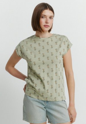 Junge Frau mit glatten braunen Haaren, die ein salbeigrünes, kurzärmeliges Oberteil mit Blumenmuster und hellblaue Jeansshorts trägt.