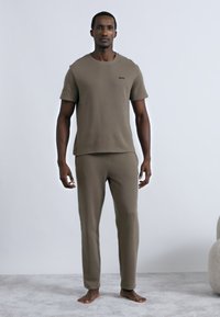 Λαδί πράσινο, εφαρμοστό βαμβακερό t-shirt με κοντά μανίκια και διακριτικό λογότυπο· ταιριαστό ελαφρώς στενό παντελόνι με ελαστική μέση· ξυπόλητος σε υφασμένο γκρι πάτωμα.