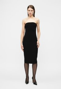 Γυναίκα που φοράει μαύρο μίντι φόρεμα χωρίς τιράντες, μαύρες διαφανείς καλσόν, μαύρες μυτερές γόβες και μακριά κρεμαστά σκουλαρίκια, στέκεται μπροστά σε ένα απλό φόντο.