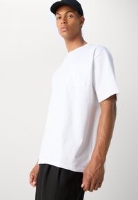Vit kortärmad t-shirt med rund halsringning och bröstficka på vänster sida, tillverkad av mjukt bomullstyg, i kombination med svarta byxor.