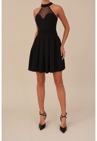 Černé halter šaty s průhledným tělem a skládanou sukní. Mají výstřih do srdce a jsou doplněny lesklými černými botami na jehlovém podpatku.