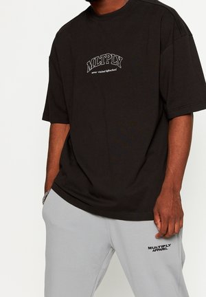 Man wearing a black oversized T-shirt with “MLTPLY never visited highschool” and light gray pants with “MULTIPLY APPAREL” text.
