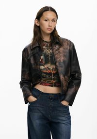 Smeđa kožna skraćena jakna s sjajnom površinom, u kombinaciji s uzorkastom majicom i opuštenim plavim trapericama s dugmetom.