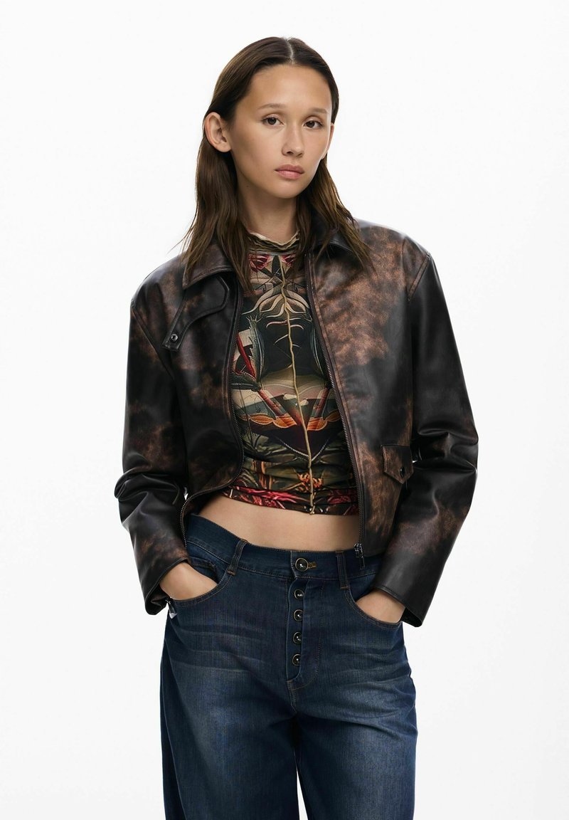 Smeđa kožna skraćena jakna s sjajnom površinom, u kombinaciji s uzorkastom majicom i opuštenim plavim trapericama s dugmetom.