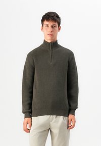 Giovane uomo che indossa un maglione a costine verde scuro con zip a quarto e pantaloni di un beige chiaro, in piedi contro uno sfondo bianco.