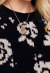 Gul hängande halsband med en oval form och graverad text, bärs över en mönstrad svart och vit stickad tröja.