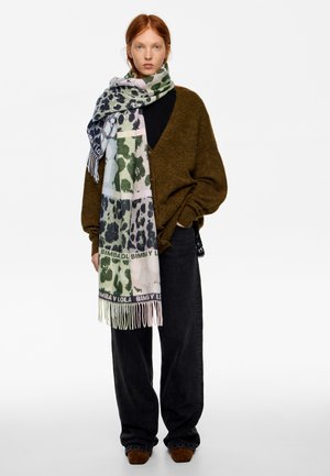 Mujer con cárdigan marrón de gran tamaño, vaqueros negros de pierna ancha, zapatos marrones y una gran bufanda multicolor con estampado de animal alrededor del cuello.