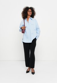 Camisa de botões azul clara com detalhes em ponto cruz, combinada com calças largas pretas e sapatos pretos. Design simples com um corte descontraído.