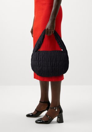 Sac à main noir matelassé avec un design en forme de U et une poignée épaisse et texturée, porté par une personne vêtue d'une robe rouge et de chaussures à talons noires.