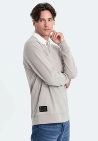 Polo grigio chiaro a maniche lunghe con colletto bianco a contrasto, tessuto texture, etichetta nera con logo sul fondo e vestibilità rilassata indossato da un modello.