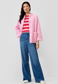 Vrouw draagt een roze, pluizig jasje met knopen, een rood-roze gestreept shirt, wijde blauwe spijkerbroek en witte sneakers, staand tegen een effen achtergrond.