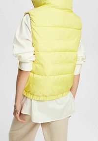 Gilet imbottito giallo con colletto alto, caratterizzato da sezioni trapuntate e orlo arruffato. Indossato sopra una blusa color crema e pantaloni beige.