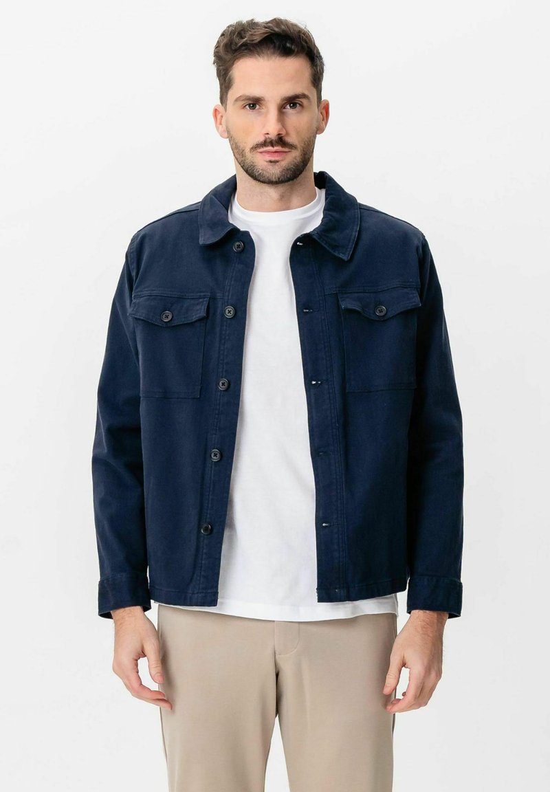 Homme portant une veste bleu marine boutonnée par-dessus un t-shirt blanc et un pantalon beige, debout devant un fond blanc uni.