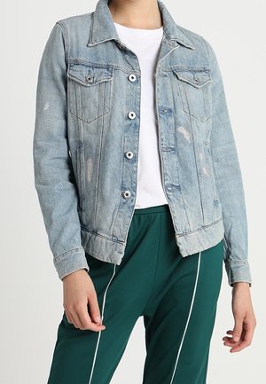 Persoon die een lichtblauwe spijkerjas draagt over een wit overhemd en donkergroene broek met witte verticale strepen.