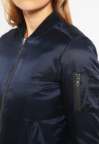 Námornícky modrá saténová bomber bunda s izolačným vreckom na ľavom rukáve a rebrovaným golierom. Hladká textúra s priliehavým strihom.