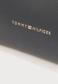 Čierna koža s textúrou a vytlačeným názvom značky "TOMMY HILFIGER" v zlatej farbe, obsahujúca obdĺžnikový akcent loga.