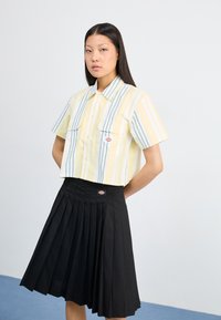 Saīsināts dzelteno un balto svītroto krekli ar divām krūšu kabatām, apvienots ar melnu svārku ar pleats, kam ir logotipa plāksteris jostasvietā.