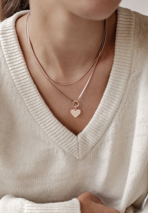 Two layered rose gold necklaces; one with a delicate chain and heart pendant, both featuring a smooth, polished finish. Worn over a cream sweater.