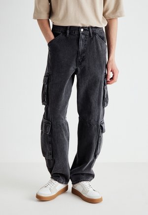 Pantaloni cargo in denim nero con molte tasche, vestibilità comoda e leggera scoloritura. Abbinati a sneakers bianche con suole marroni chiari.