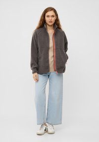 Giacca in fleece grigia con colletto alto e zip arancione, indossata sopra un top beige. In abbinamento a jeans a gamba larga azzurri chiari e sneakers bianche.