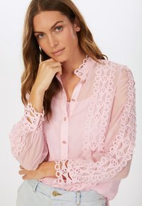 Helle rosa Bluse mit Knopfverschluss und intricater Spitzenverzierung an den Ärmeln. Transparenter Stoff mit bestickten Mustern. Rundhalsausschnitt.