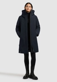 Lang marineblå dunjakke med hætte, frontlynlås og to sidelommer. Båret over et sort outfit, parret med sorte kraftige støvler.