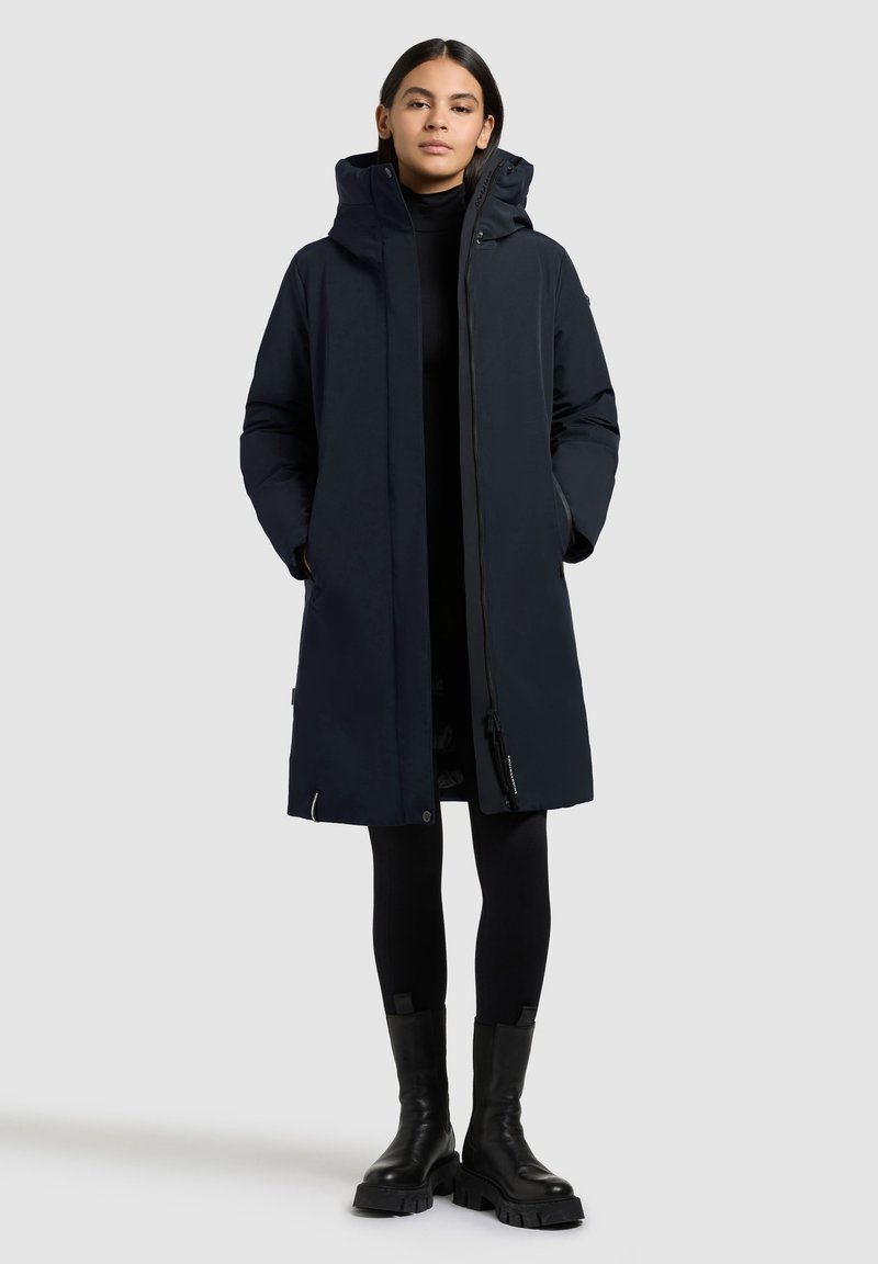 Lang marineblå dunjakke med hætte, frontlynlås og to sidelommer. Båret over et sort outfit, parret med sorte kraftige støvler.