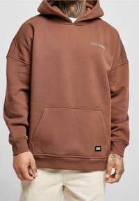 Sudadera marrón con un bolsillo frontal, capucha y puños acanalados. Presenta un bordado de logo mínimo. Tejido suave con una textura lisa.