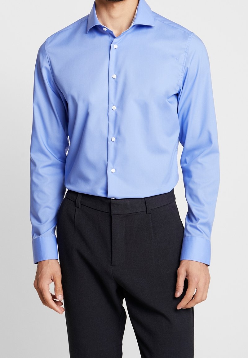 Ljusblå skjorta med knäppning och strukturerad krage, långärmad, i kombination med mörka skräddarsydda byxor. Slät tyg med en lätt glans.