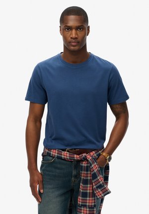 Camiseta de manga corta azul marino de algodón, combinada con unos jeans de mezclilla oscuros. Una camisa de franela a cuadros está atada a la cintura.