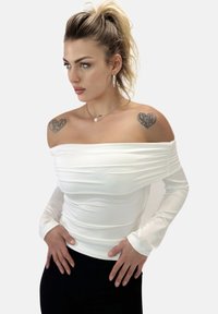 Top bianco aderente con scollatura off-the-shoulder e maniche lunghe; presenta una texture arricciata e un tessuto liscio. Il modello lo indossa con pantaloni neri.