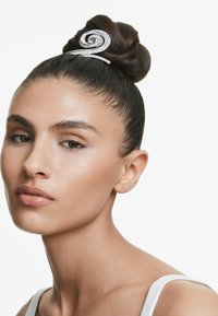 Un mannequin regarde la caméra avec confiance, affichant un chignon élégant agrémenté d'un accessoire décoratif, sur fond blanc.