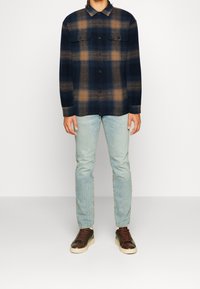 Marineblauwe geruite shirt met bruine accenten, knoopsluiting, twee borstzakken, gecombineerd met lichtblauwe jeans en bruine sneakers met crèmekleurige zolen.