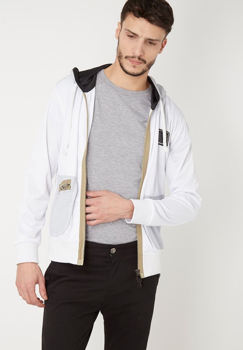 Sudadera blanca con cremallera y capucha negra. Presenta un bolsillo gris claro y una cremallera dorada. Combinada con una camiseta gris y pantalones negros.