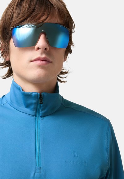 Modrý svetr se čtvrtým zipem a hladkou texturou, na hrudi malý logo. Celkový vzhled doplňují velké modré zrcadlové sluneční brýle.