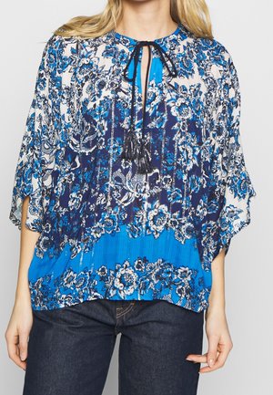 Losse blauwe en witte blouse met bloemenprint en zwarte tasselveters bij de hals, gedragen met donkerblauwe spijkerbroek, zichtbaar van schouders tot heupen.