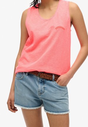 Personne portant un top sans manches corail rose avec le texte "SUPRADRY" et un short en jean bleu clair avec une ceinture tressée marron, une main dans la poche.