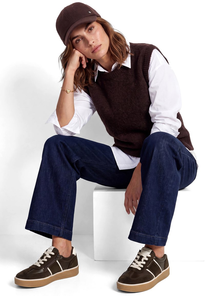 Jeune femme portant une casquette marron, un gilet foncé sur une chemise blanche, un jean bleu et des baskets marron, assise sur un cube blanc la main sur le visage.