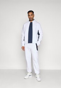 Biały sportowy dres z granatowymi akcentami. Kurtka na zamek, koszulka z okrągłym dekoltem i dopasowane spodnie. Gładki materiał, minimalistyczny design, luźny krój.