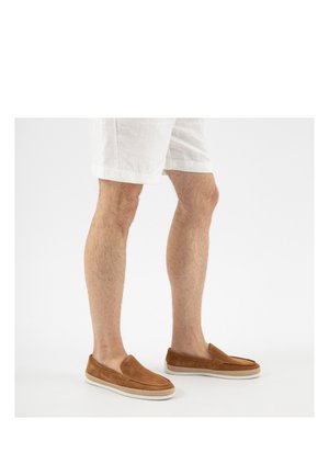 Gambe che indossano pantaloncini bianchi e scarpe slip-on in camoscio color cuoio con suole di gomma bianche, in piedi su uno sfondo bianco semplice.