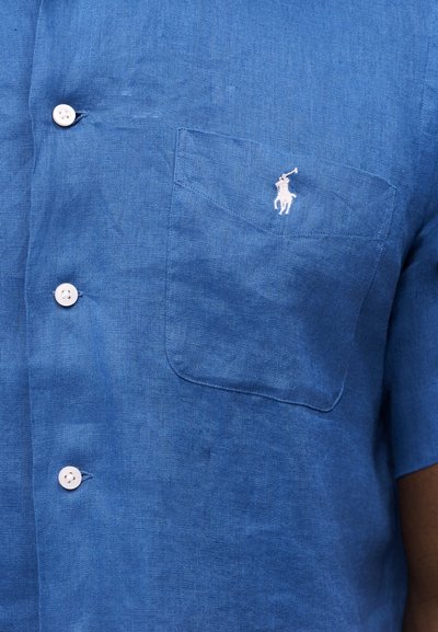 Zils linu īss piedurkņu krekls ar baltām pogām un krūšu kabatu, uz kura redzams mazs balts zirgs ar jātnieku.