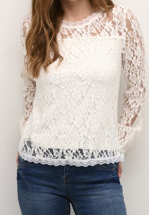 Haut à manches longues en dentelle blanche avec un motif floral, décolleté festonné et bord en dentelle au bas. Associé à un jean en denim bleu.
