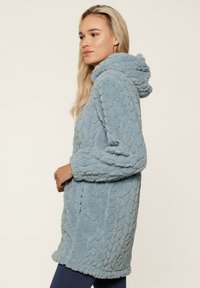 Veste extérieure à capuche bleu clair en tissu doux et texturé avec un motif en scaille en relief. Dotée de manches longues et de poches latérales.