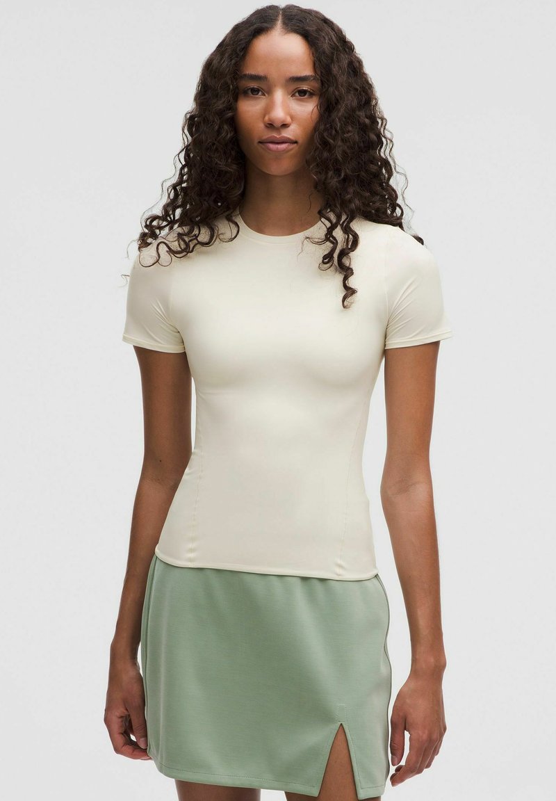 Top beige chiaro a maniche corte con un design aderente. Abbinato a una gonna verde chiaro con uno spacco su un lato. Tessuto dalla consistenza liscia.