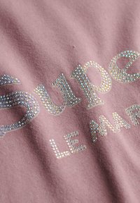 Rosa stof med ordene "Super" og "LE MAR" udsmykket med skinnende, multikolorede rhinsten i en dekorativ skrifttype.